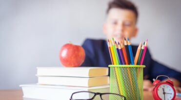 Schoolboy smiling behind a desk with books, pencils, and an alarm clock symbolizing study and creativity.